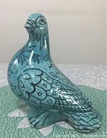 Front and side view of turquoise glazed ceramic bird figurine with black painted feather and facial details on a green textured mat.