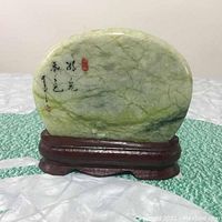 Front view of the oval jade sculpture showing green and white veining, black Chinese calligraphy, and a red seal on a wooden stand.