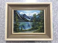 Framed oil painting on board depicting a mountain lake landscape with trees and snow-capped mountains.