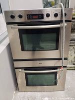 Front view of stainless steel double oven showing both doors closed, control panel, and power cable.