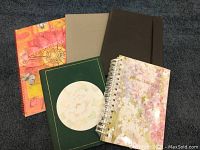 Photo showing four notebooks and one spiral-bound phone/address book with floral cover arranged on dark carpet.