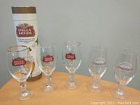 Five beer glasses displayed on table with package behind. Three Stella Artois glasses with gold rims and logos, two Innis & Gunn glasses with white and red logos.