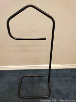 Black metal garment holder stand with rectangular base and a curved arm for hanging clothes, shown against a wall and carpeted floor.