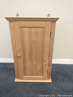 Front view of pine shelving unit with closed door, showing wood grain and door knob, with two wall hooks on top for hanging.