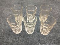Set of six large clear glass drinking glasses arranged in a group on carpet, showing their consistent shape and design.