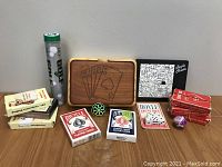Photo showing a variety of games including multiple decks of cards from Bicycle and Hoyle, dice, a golf game in a tube, a puzzle box with black and white pattern and wooden carrying case for cards/games.