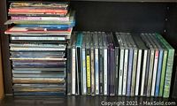 Photo displaying two stacks of music CDs arranged on a black surface; left stack larger with mixed case colors, right stack neatly lined with readable spines.