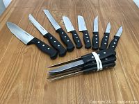 Set of 8 assorted Farberware cutting knives and a set of 6 steak knives with black handles and riveted tangs on a wooden surface