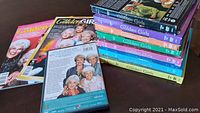 Seven colorful DVD boxed sets of The Golden Girls seasons stacked on a table alongside commemorative magazines and an additional DVD about the cast.