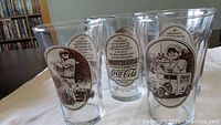 Six clear glass Mother's Pizza Coca-Cola collectible glasses with vintage brown illustrations and text, shown standing grouped on a table.