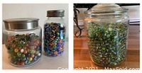 Three jars containing assorted glass marbles and flat colored stones. Two jars have a modern wide-mouth design with metal lids, one smaller, one larger. The third jar is a larger rounded glass jar with a glass lid containing many green-tone glass marbles.
