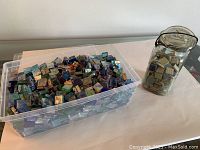 Large clear plastic container filled with small assorted glass tiles next to mason jar filled with small glass tiles.