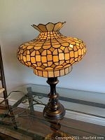 Full view of stained glass lamp on glass table showing golden-hued patterned glass shade and dark metal base.