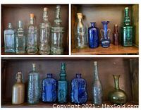 Group of 17 assorted glass bottles displayed on wooden shelves, showing variety of sizes, shapes, and colors including clear, green, blue, amber, and bronze-tinted bottles with some having corks or closures.