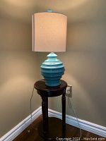 Photo of turquoise-green ceramic ribbed lamp on small round dark wood side table against wall, beige shade visible with some damage.