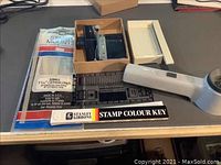 Photo shows the Stanley Gibbons Stamp Colour Key, stamp mounts packaging, perforation gauge, stamp cutter, and the white magnifying glass with light all arranged on a gray surface.