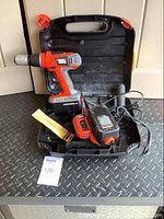 Photo showing the cordless drill with battery attached, charger, and black plastic carrying case on the floor.