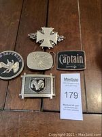 Top view of five belt buckles laid on wooden surface, including Led Zeppelin, Dodge, Captain Morgan, Iron Cross, and weed leaf designs.
