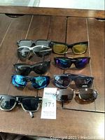 Nine pairs of sunglasses arranged on wooden surface showing various frame styles and lens colors.
