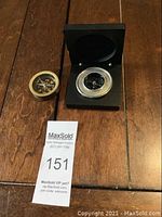 Two compasses on wooden floor, one brass with black face compass, one silver compass in black display box