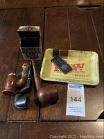 Photo of three tobacco pipes, wooden cigarette box, RAW ashtray, and cigar cutter arranged on wooden table