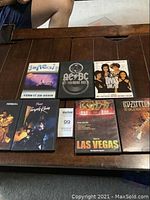 Photo showing all 7 concert DVDs on a wooden surface, including Genesis, AC/DC, Divas Live 99, Prince (Purple Rain), Kiss, and Led Zeppelin titles.