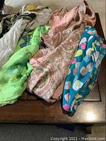 Overview of several women's shawls and scarves displayed on a wooden table showing various colors and patterns including green, pink paisley, and blue floral design.