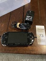 Front view of black Sony PSP-1001 handheld console with screen and buttons, power adapter with cord next to it.