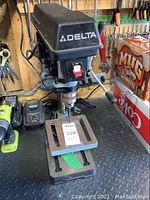 Front angled view of Delta drill press showing brand label, on/off switch, drill chuck and handle, metal base with slotted table.
