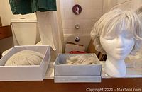Three wigs shown, one on styrofoam head and two stored in boxes on bathroom counter near toilet and shower curtain.