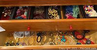 Top drawer containing assorted costume necklaces, bracelets, earrings, and pins, sorted in compartments and loosely placed.