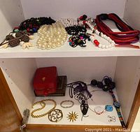 Two shelves showing an assortment of costume necklaces, bracelet, pins and two decorative boxes including a red jewelry box.