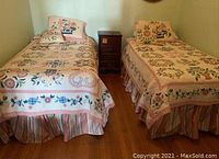 Two twin beds made up with floral quilts, pillows, shams, and striped bed skirts, arranged side by side with a small wooden nightstand between them.