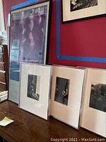 Three black and white framed photographs on a table with a large framed poster leaning against the wall.