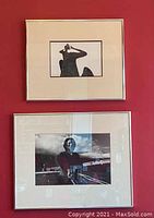 Two framed black and white photographs hung on a red wall. The upper photograph shows a person with arm raised shielding eyes, the lower photograph is a portrait with a background including text.