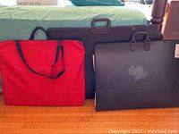Three portfolios side by side on floor: one red soft case bag, two black hard cases with handles.