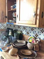 Wooden bowls, platters, woven plates, assorted mugs and glasses displayed on a cabinet top with tiled backsplash showing floral patterns.