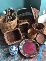 Photo of various woven baskets of different sizes and styles, ceramic plate, and ceramic ginger jar on carpeted floor.