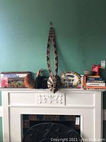Full view of mantel showing items: decorative mask, ceramic plate, figurines, books, and CD cases, arranged on a white mantel against turquoise wall