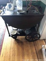 Full view of the tea cart showing drop leaf top with glass, wooden legs, large metal wheels, and lower shelf with metal plates and container.