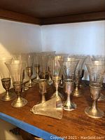 Overall view of all 12 silver base wine glass holders with clear etched glass inserts on wooden shelf, one insert lying on its side