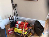 Photo of fireplace tool set including poker, tongs, broom, shovel with wooden handles next to a basket containing Duraflame firelogs and fire starters, and a black metal ash bucket.