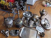 Full assortment of silver plate items on hardwood floor including candelabra, trays, coffee pot, covered dish, and candle holders showing tarnish and need for cleaning.