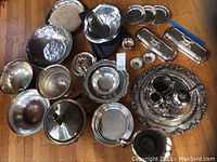 Photo showing a variety of silver plated trays, bowls, plates, a pitcher, and a coffee service set arranged on wooden floor. Items display tarnish and signs of age.