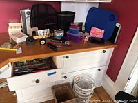 Photo of the office supplies spread on a wooden desk and inside open drawers, showing lap desk, push pins, paper punch, rolodex cards, pencils and pens, small containers, black clock, paper weight.