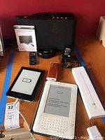 Photo of all items together on wooden desk including two Kindles, Apple Pencil box, Gigaware docking speaker, power adapter, remote controller, and USB flash drive