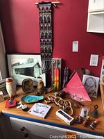Wide view of the lot showing worry dolls, dream catcher, bracelets, framed art, bamboo gavel, hanging tapestry, and various decorative items on a wooden dresser against a red wall.