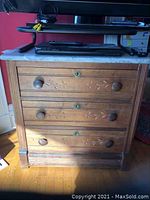 Front view of wooden chest of drawers with marble top resting on it, showing three drawers with round knobs and floral carvings.