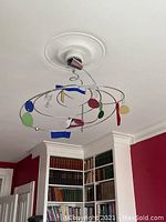 Ceiling fixture mounted and shown from a side angle in a room with red walls and bookshelves, showing colorful abstract hanging shapes.