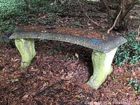Wide view showing the entire curved bench with cement top and two supporting decorative legs outdoors on leafy ground, showing weathering and debris on top surface.